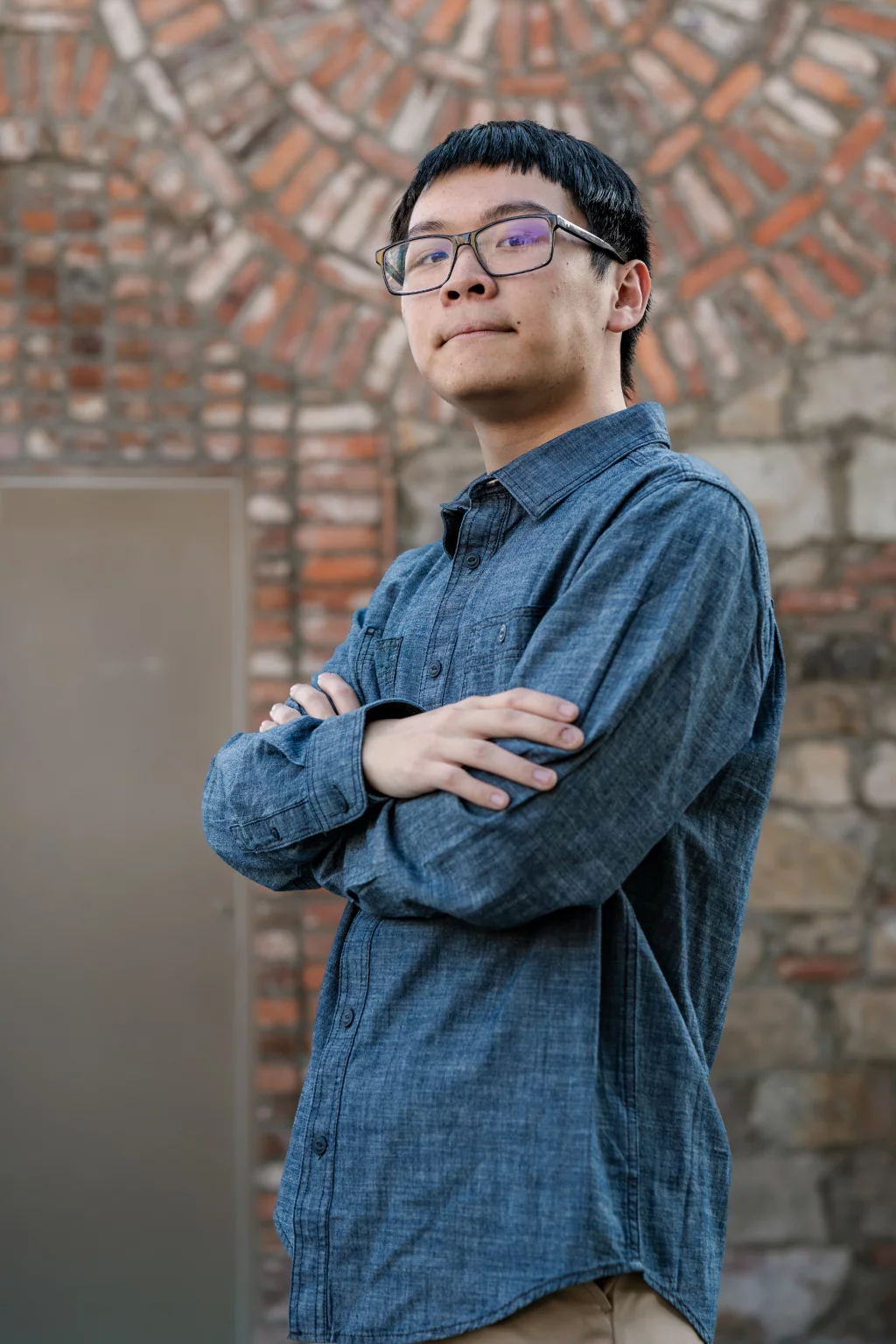 Johnny's senior photos taken at BridgeWay Station, Simpsonville, SC: Young person with short dark hair and glasses stands with arms crossed, wearing a blue button-up shirt, in front of a stone and brick wall.