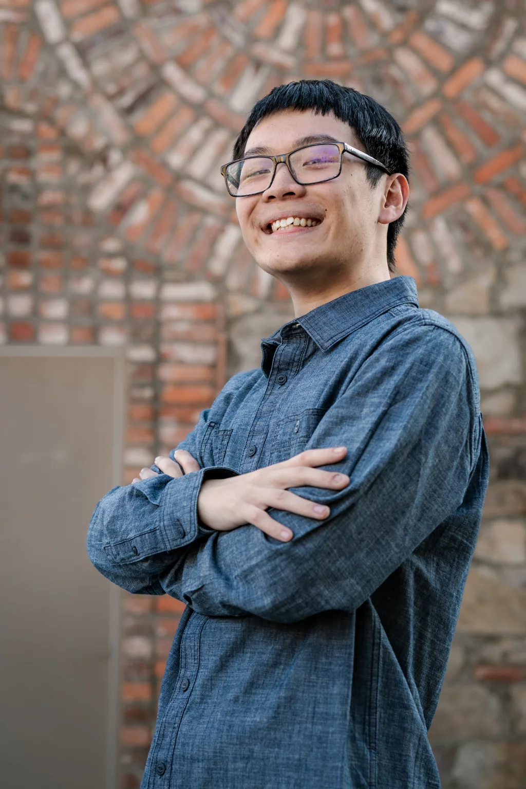 Johnny's senior photos taken at BridgeWay Station, Simpsonville, SC: Young person with short black hair and glasses smiling with arms crossed, wearing a blue button-up shirt, standing in front of a brick and stone wall.