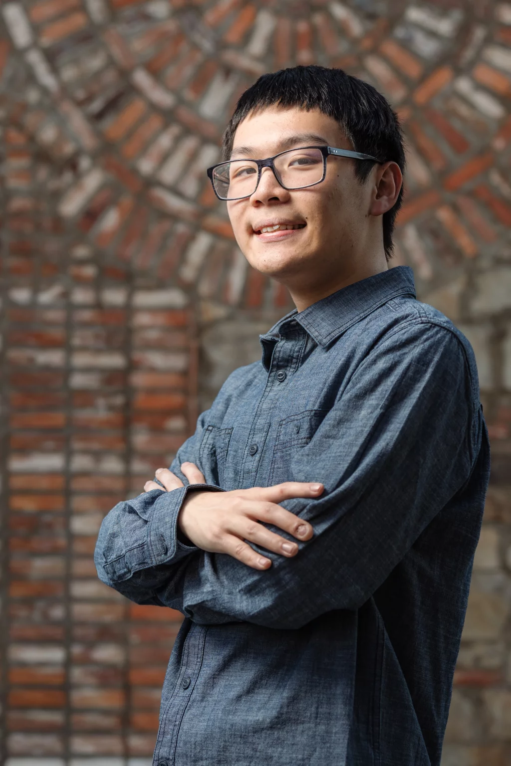 Johnny's senior photos taken at BridgeWay Station, Simpsonville, SC: Young man with short dark hair and glasses, wearing a blue button-up shirt, standing with arms crossed and smiling in front of a brick and stone wall.