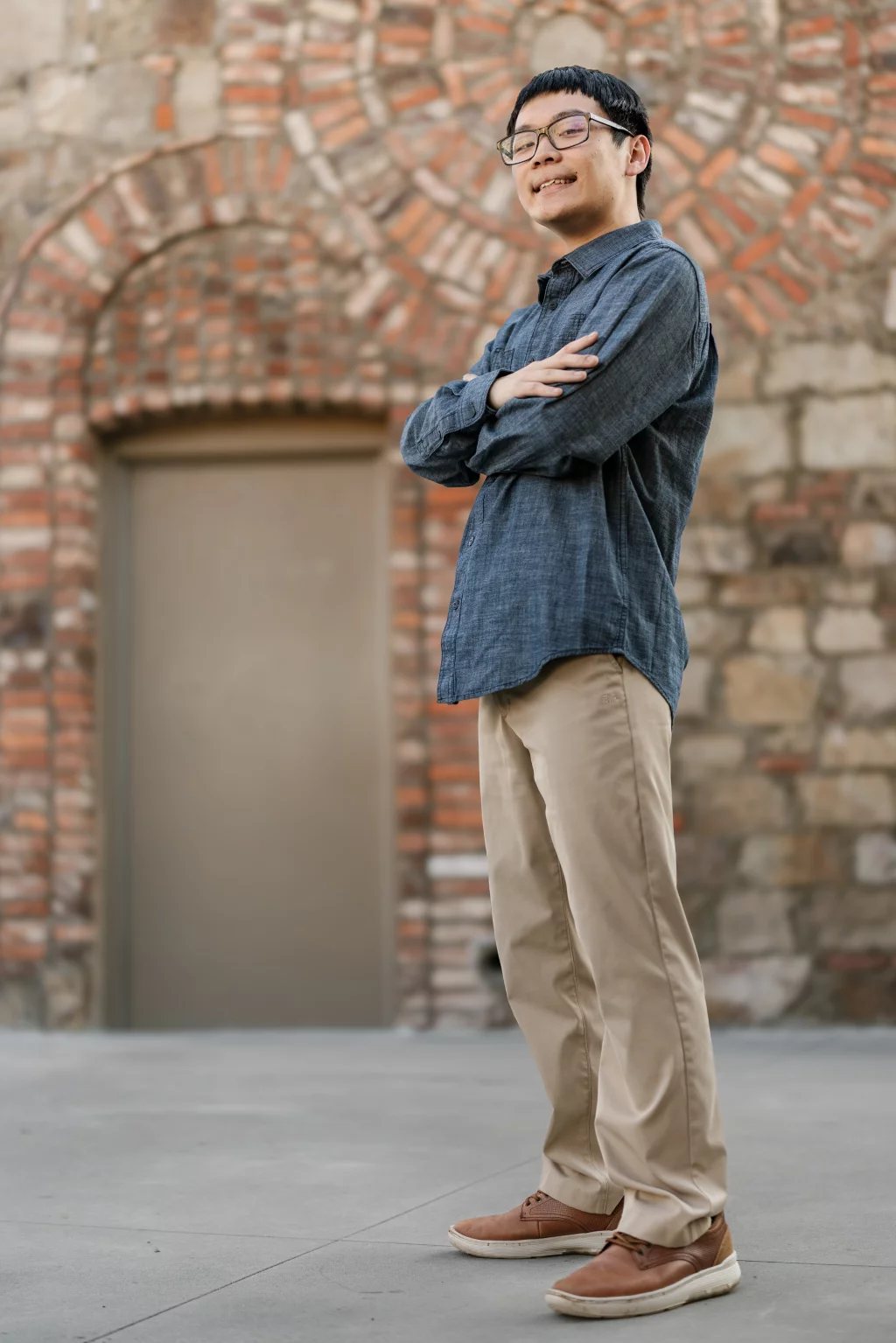 Johnny's senior photos taken at BridgeWay Station, Simpsonville, SC: A young man stands outdoors in front of a brick and stone wall with an arched doorway, smiling with arms crossed, wearing glasses, a blue button-up shirt, khaki pants, and brown shoes.