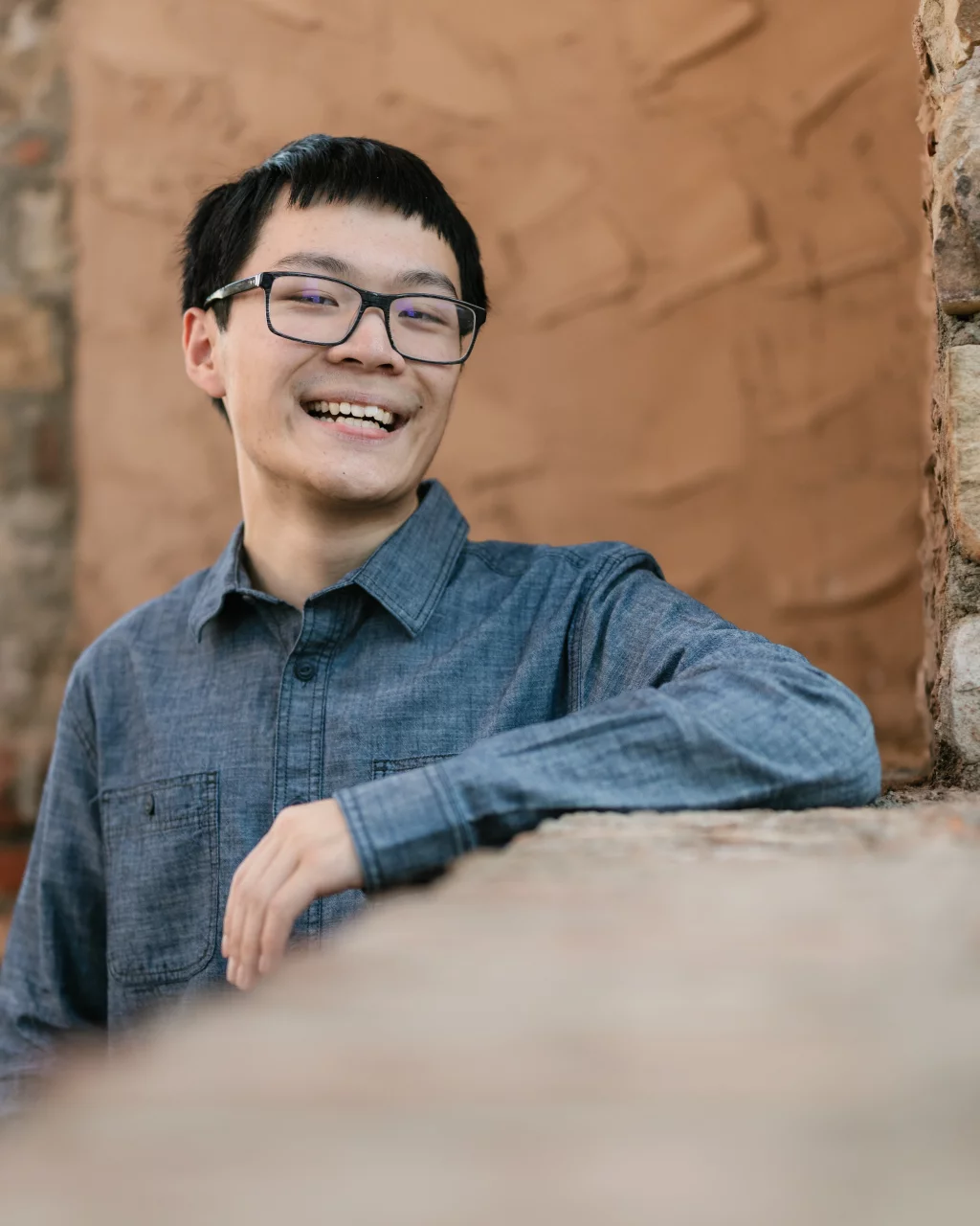 Johnny's senior photos taken at BridgeWay Station, Simpsonville, SC: A young man with short black hair and glasses smiles while leaning on a stone ledge, wearing a blue button-up shirt. The background is a textured tan wall with stone accents.