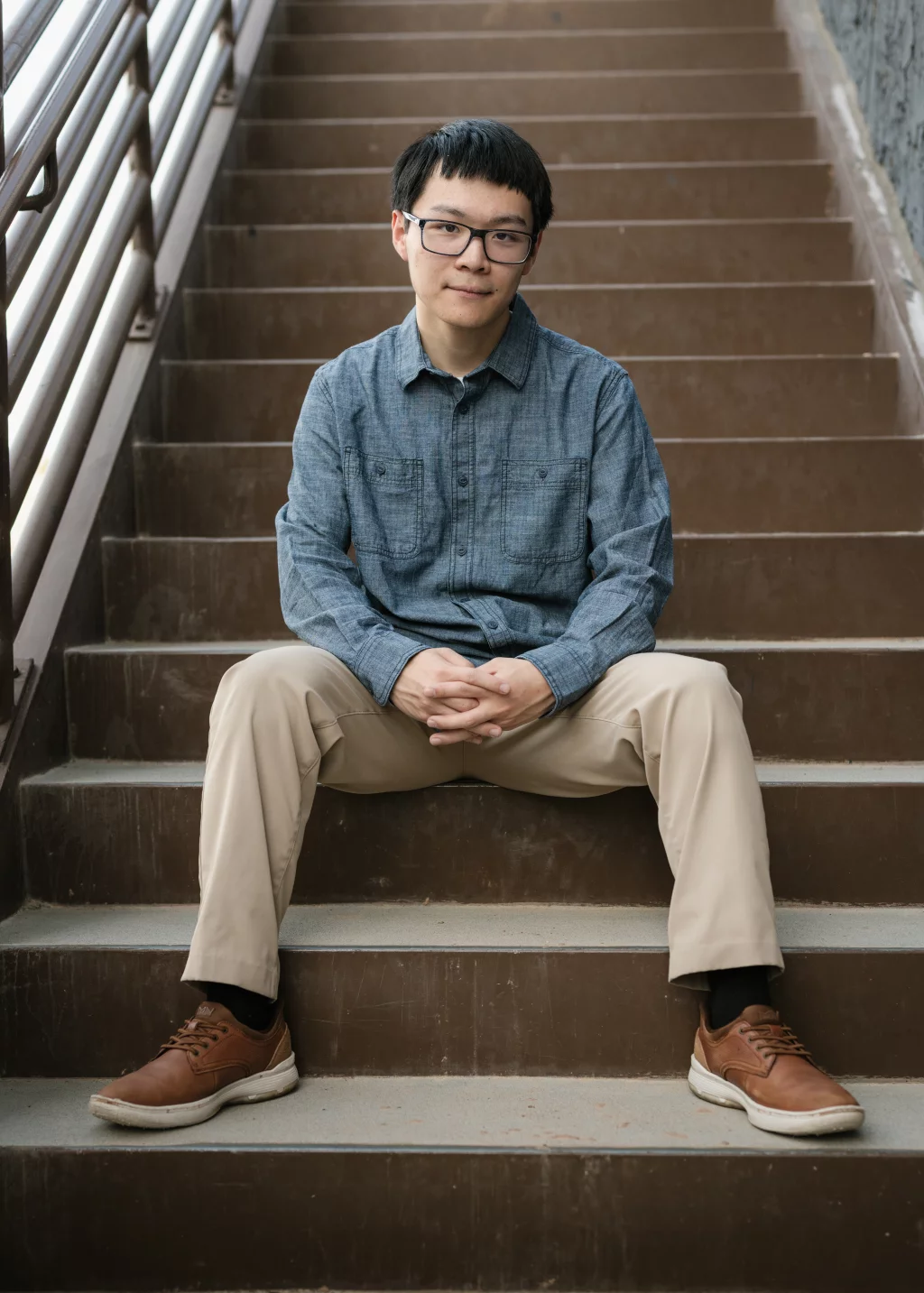 Johnny's senior photos taken at BridgeWay Station, Simpsonville, SC: A young man with short black hair and glasses sits on outdoor steps, wearing a blue button-up shirt, khaki pants, and brown shoes, with hands clasped in his lap.