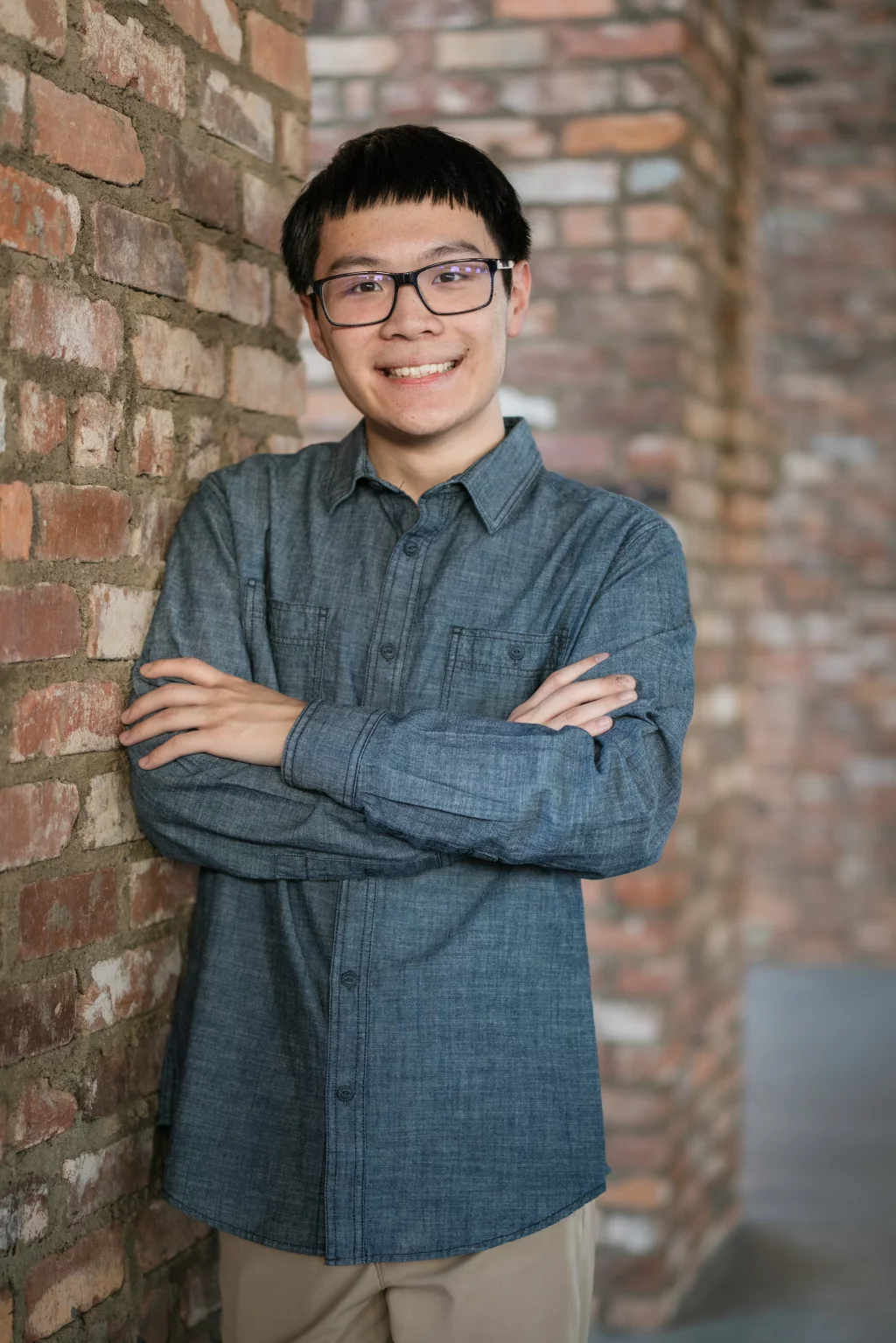 Johnny's senior photos taken at BridgeWay Station, Simpsonville, SC: Young person with short dark hair and glasses, smiling with arms crossed, standing against a brick wall.