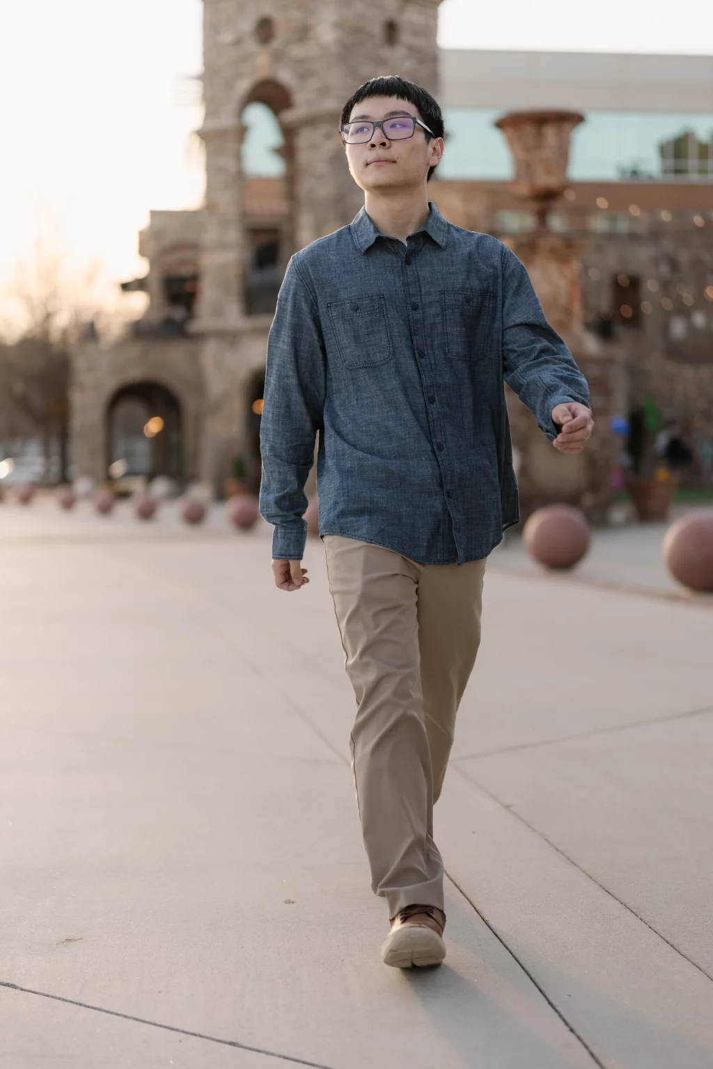 Johnny's senior photos taken at BridgeWay Station, Simpsonville, SC: A young man wearing glasses, a blue button-up shirt, and khaki pants walks confidently on a paved path with a stone building and round planters in the background.