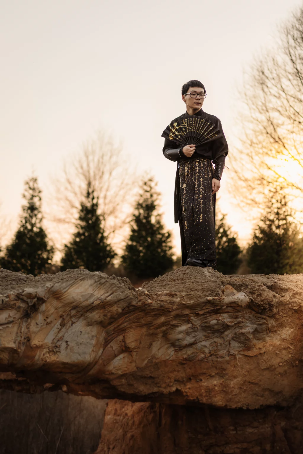 Johnny's senior photos taken at BridgeWay Station, Simpsonville, SC: A person in a black and gold robe holding a decorative fan stands on a rocky ledge outdoors at sunset, with trees in the background.