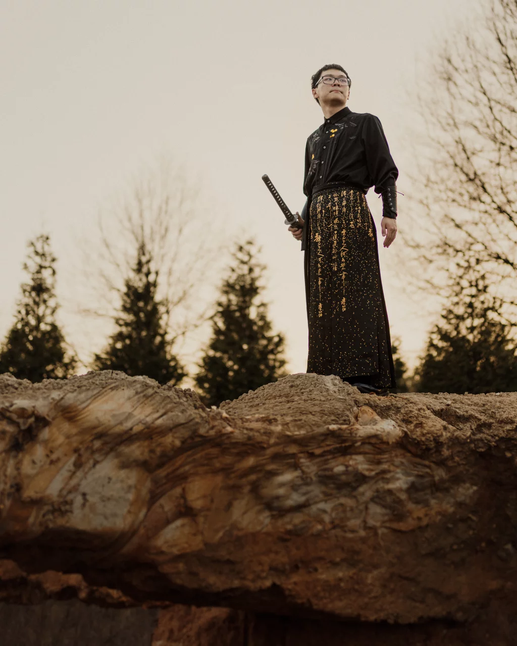 Johnny's senior photos taken at BridgeWay Station, Simpsonville, SC: A person in a black and gold outfit stands confidently on a large rock, holding a sword, with trees and a cloudy sky in the background.