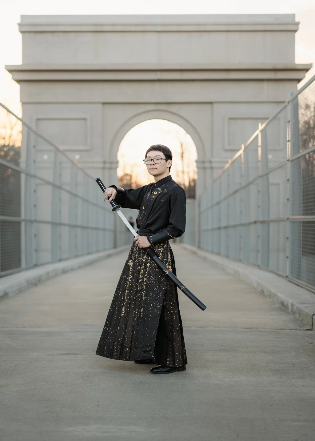 Johnny's senior photos taken at BridgeWay Station, Simpsonville, SC: A person wearing a black and gold-patterned robe stands on a pedestrian bridge, holding a katana sword, with a large stone archway in the background at sunset.