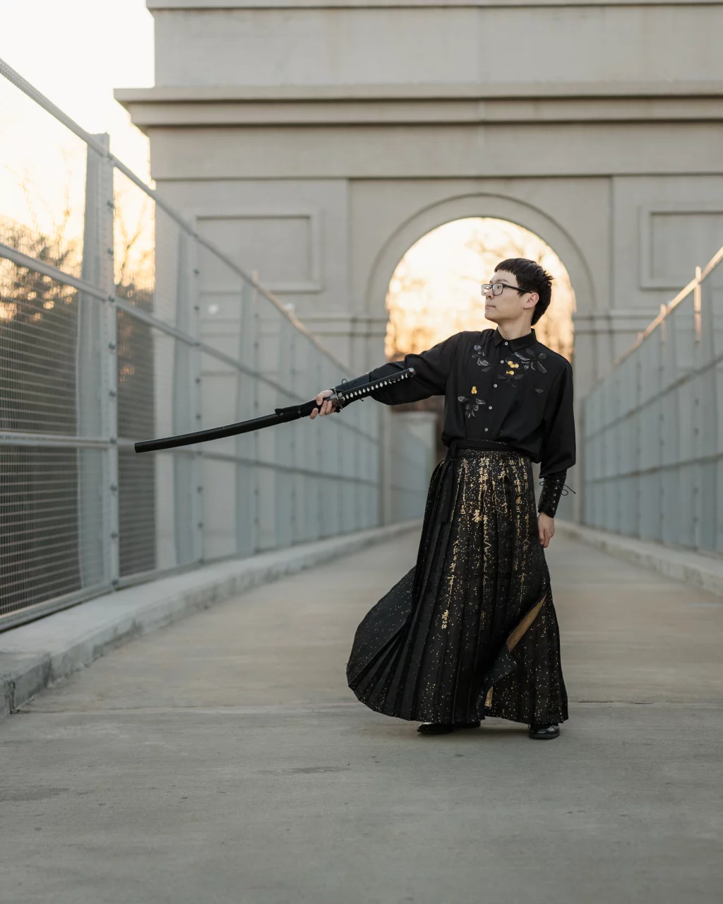 Johnny's senior photos taken at BridgeWay Station, Simpsonville, SC: Person wearing a black shirt and a long black and gold skirt stands on a walkway, holding a sheathed sword horizontally, with a large stone archway in the background.