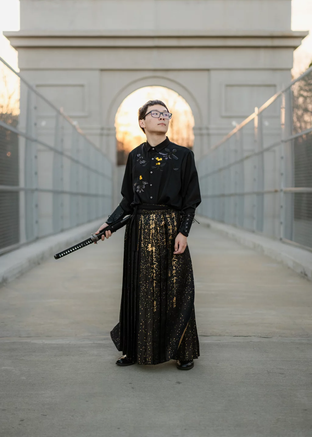 Johnny's senior photos taken at BridgeWay Station, Simpsonville, SC: A person stands on a pedestrian bridge at sunset, wearing a black shirt with embroidered flowers and a long, black and gold patterned skirt, holding a sheathed katana. An archway and metal railings frame the background.