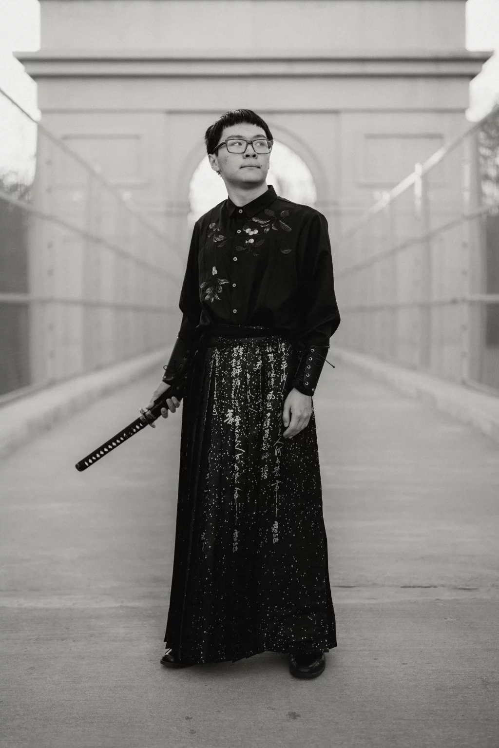 Johnny's senior photos taken at BridgeWay Station, Simpsonville, SC: Black and white photo of a person standing on a bridge, wearing glasses and a long, dark outfit with floral and calligraphy patterns, holding a sheathed sword at their side.