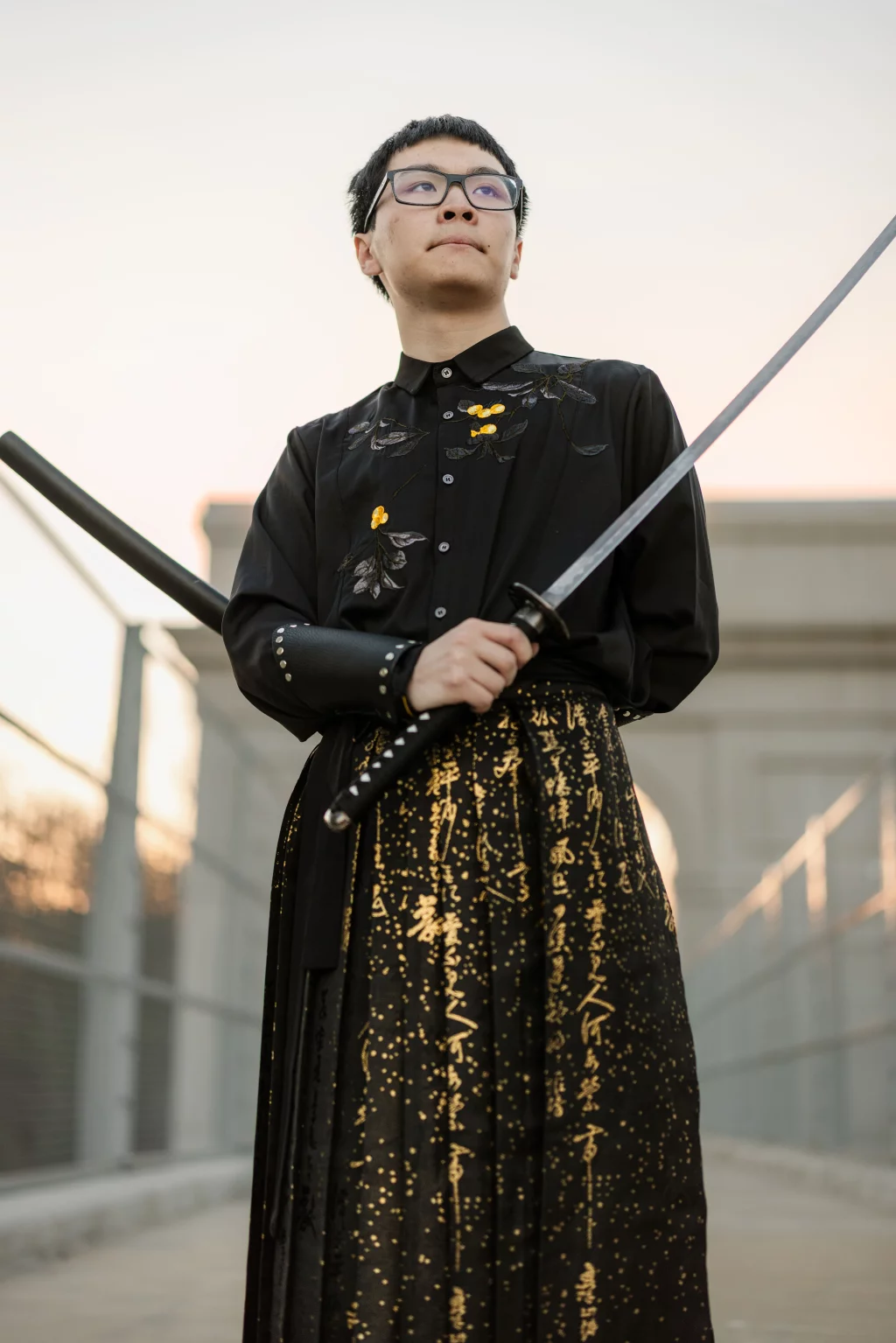 Johnny's senior photos taken at BridgeWay Station, Simpsonville, SC: A person in glasses wearing black clothing with gold embroidery and holding a katana sword stands confidently on a bridge at sunset.