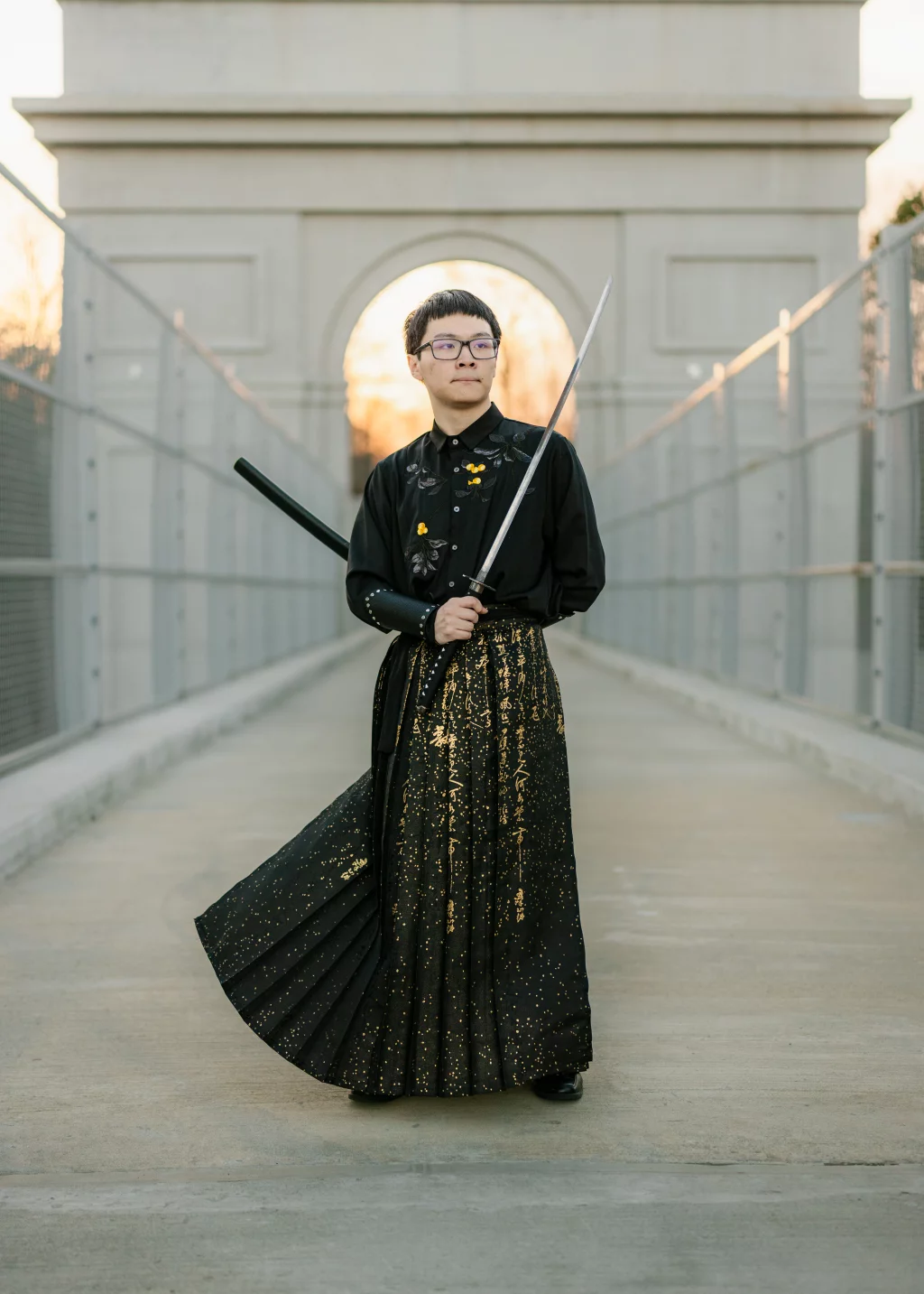 Johnny's senior photos taken at BridgeWay Station, Simpsonville, SC: Person standing on a bridge wearing a black outfit with gold designs and holding a katana sword, with an arched structure in the background.