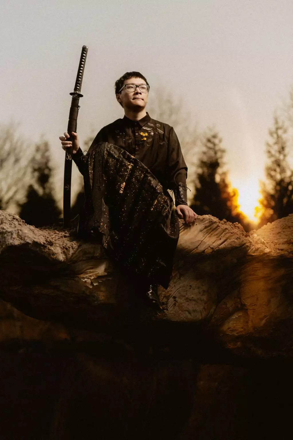 : Person wearing black and gold traditional clothing sits on a large rock, holding a sheathed sword upright, with trees and a setting sun in the background.