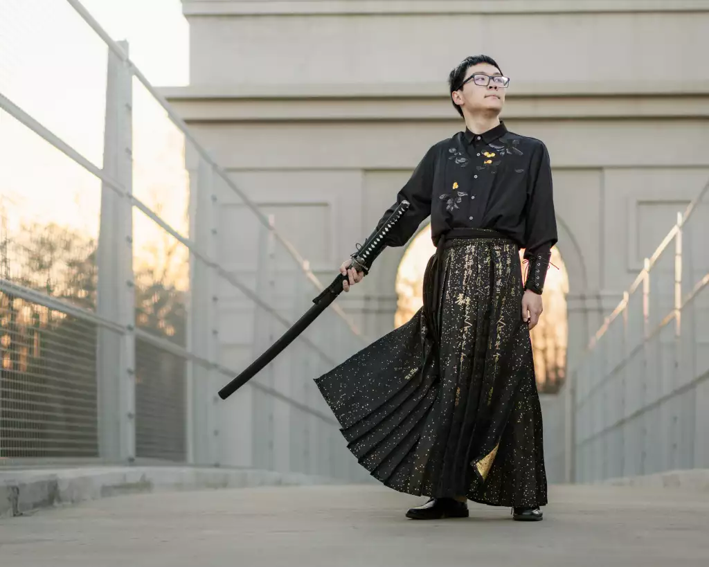 Johnny's senior photos taken at BridgeWay Station, Simpsonville, SC: Person standing confidently on a bridge, wearing traditional-inspired black and gold clothing, holding a sheathed sword, with architectural structure and sunset in the background.