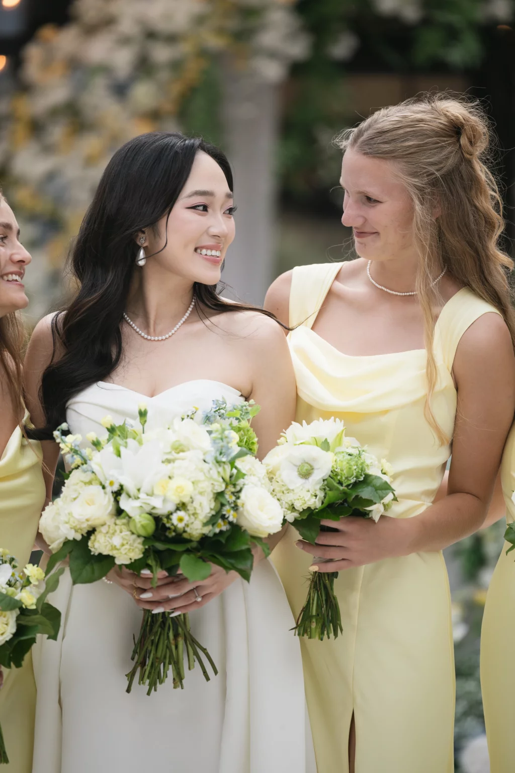 Robbie & Hannah's Wedding @ Edinburgh West, Taylors SC: Bride in a white gown and bridesmaid in pale yellow smile together, holding bouquets at an outdoor wedding.