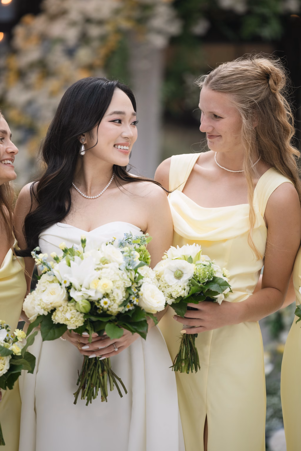 Robbie & Hannah's Wedding @ Edinburgh West, Taylors SC: Bride in a white gown and bridesmaid in pale yellow smile together, holding bouquets at an outdoor wedding.