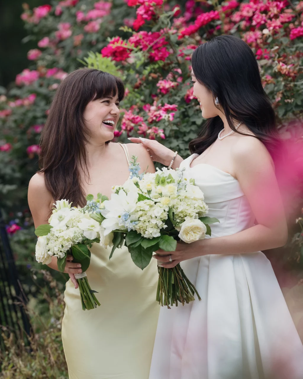 Robbie & Hannah's Wedding @ Edinburgh West, Taylors SC: Two brides laugh together while holding white flower bouquets in front of blooming pink flowers.