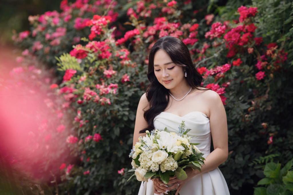 Robbie & Hannah's Wedding @ Edinburgh West, Taylors SC: Bride in a white strapless dress holding a bouquet of white flowers, posed before blooming pink roses.