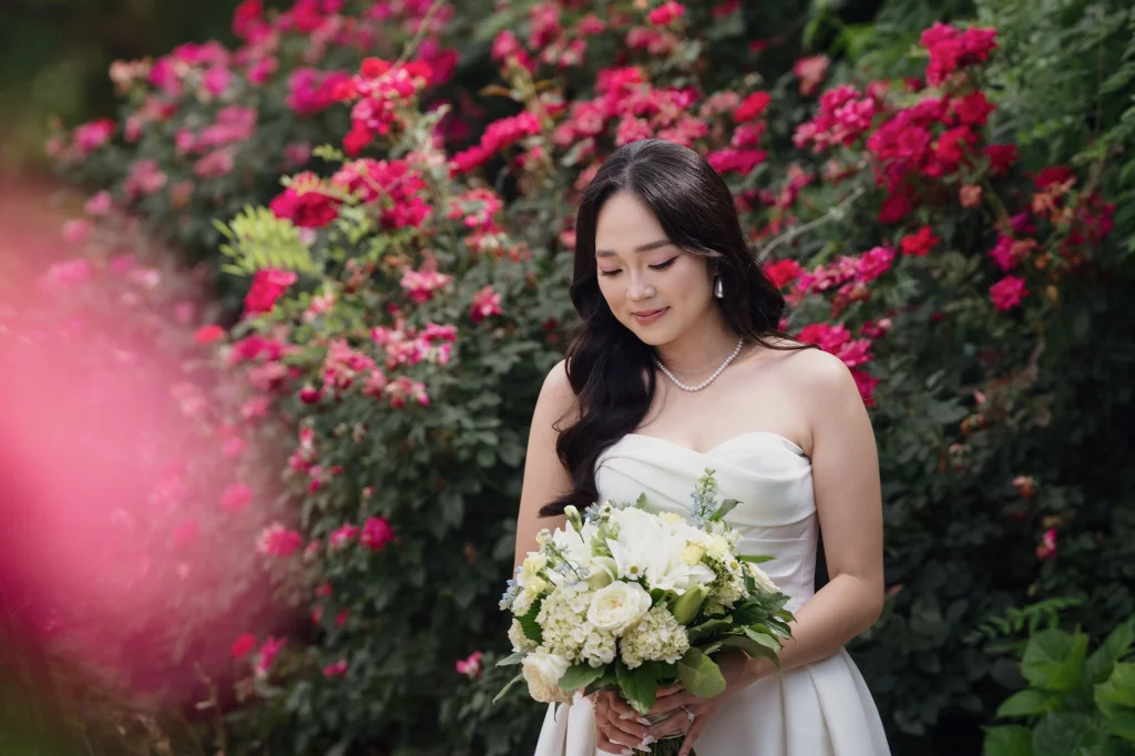 Robbie & Hannah's Wedding @ Edinburgh West, Taylors SC: Bride in a white strapless dress holding a bouquet of white flowers, posed before blooming pink roses.