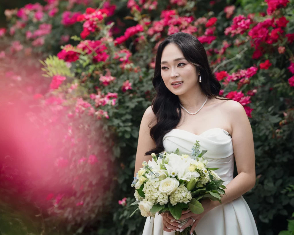 Robbie & Hannah's Wedding @ Edinburgh West, Taylors SC: Bride in a strapless white gown holding a white floral bouquet, posed before blooming pink roses.