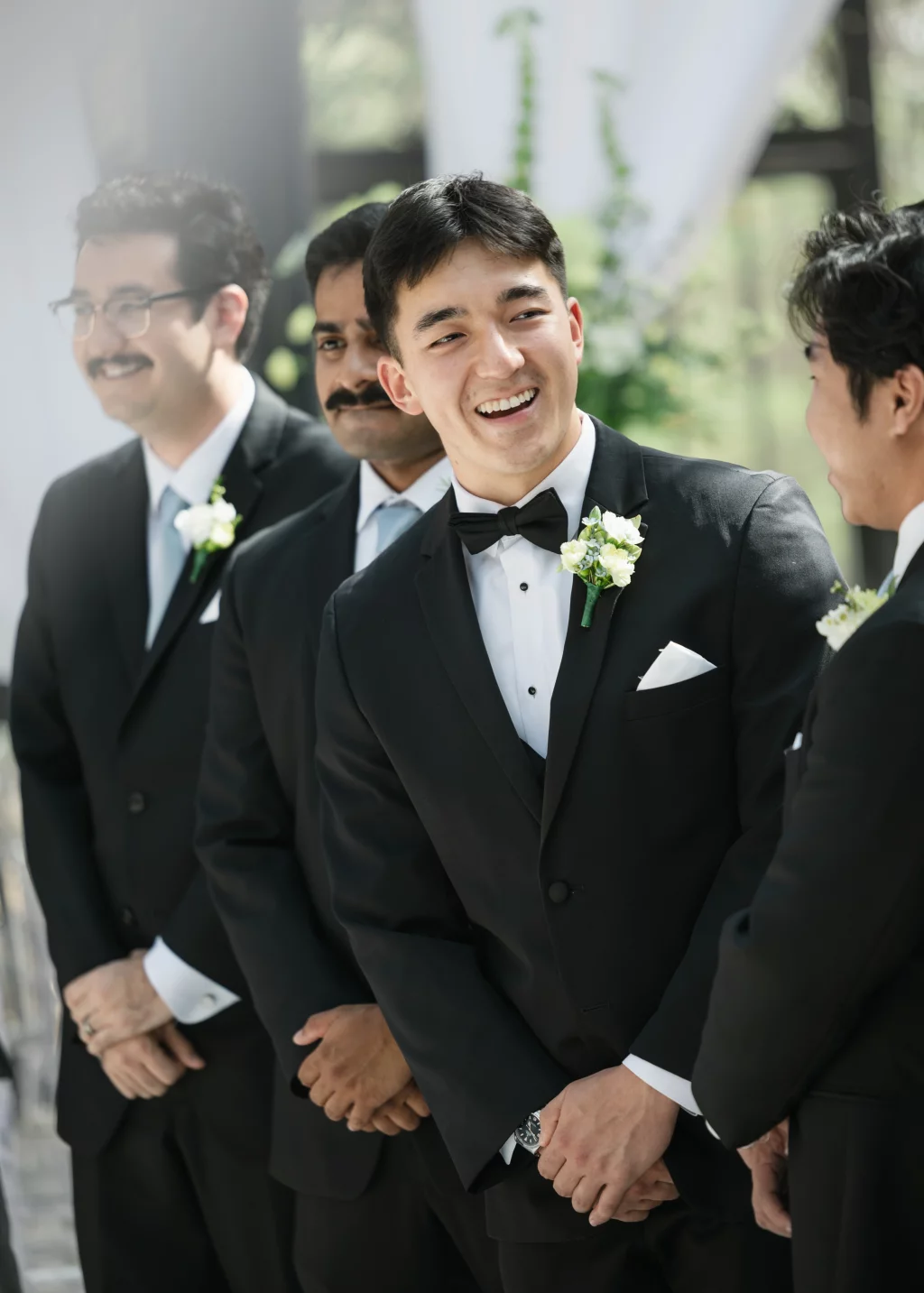Robbie & Hannah's Wedding @ Edinburgh West, Taylors SC: Smiling groom in a black tuxedo and boutonniere stands with three groomsmen outdoors.