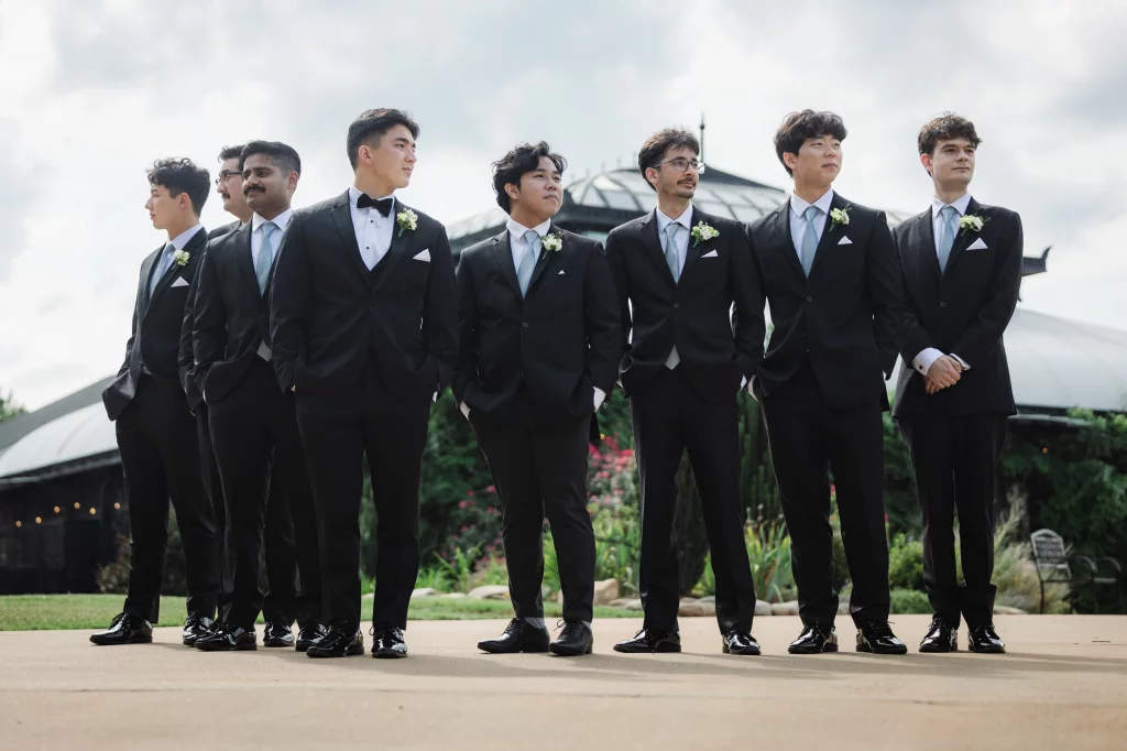 Robbie & Hannah's Wedding @ Edinburgh West, Taylors SC: Seven groomsmen in black suits and boutonnieres pose outdoors at a wedding venue.