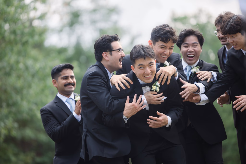 Robbie & Hannah's Wedding @ Edinburgh West, Taylors SC: Groom in a tuxedo laughs as six groomsmen playfully embrace him outdoors.