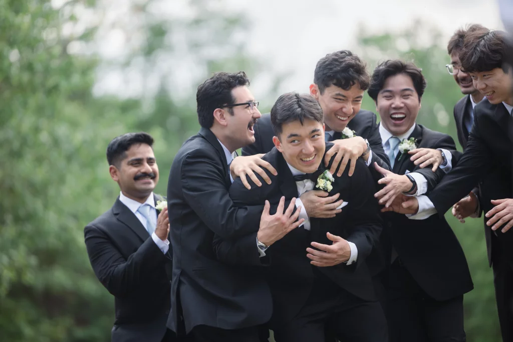 Robbie & Hannah's Wedding @ Edinburgh West, Taylors SC: Groom in a tuxedo laughs as six groomsmen playfully embrace him outdoors.