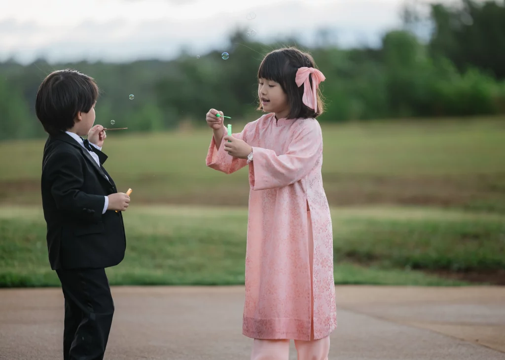 Robbie & Hannah's Wedding @ Edinburgh West, Taylors SC: Two children in formal attire blow bubbles outdoors; a boy wears a black suit, and a girl wears a pink dress with a bow.
