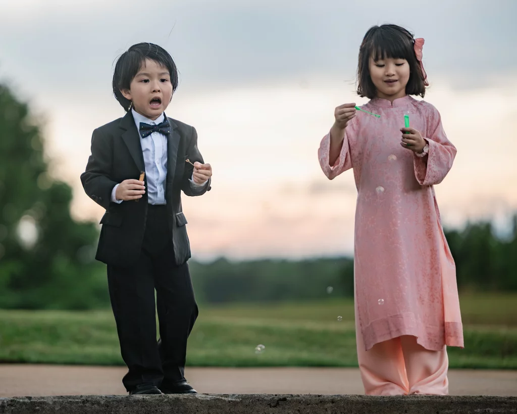 Robbie & Hannah's Wedding @ Edinburgh West, Taylors SC: Two children in formal attire blow bubbles outdoors at sunset, with the boy in a suit and the girl in a pink dress.