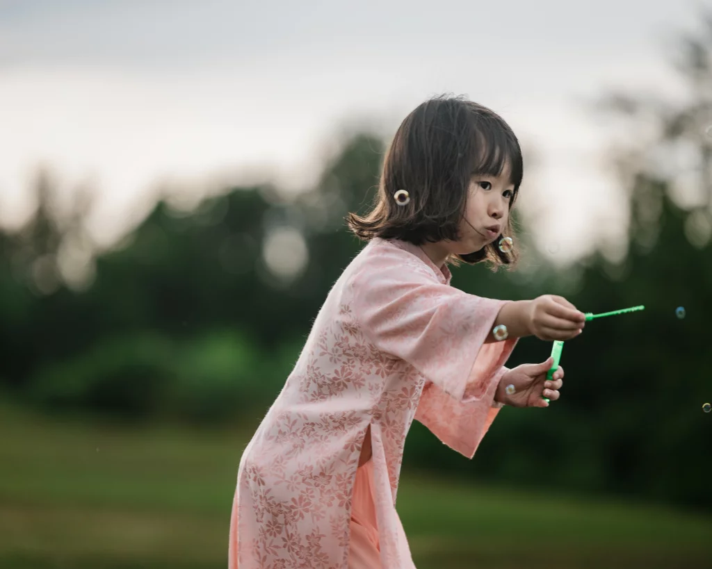 Robbie & Hannah's Wedding @ Edinburgh West, Taylors SC: Child in a pink floral dress holds a bubble wand outdoors as bubbles float around her.