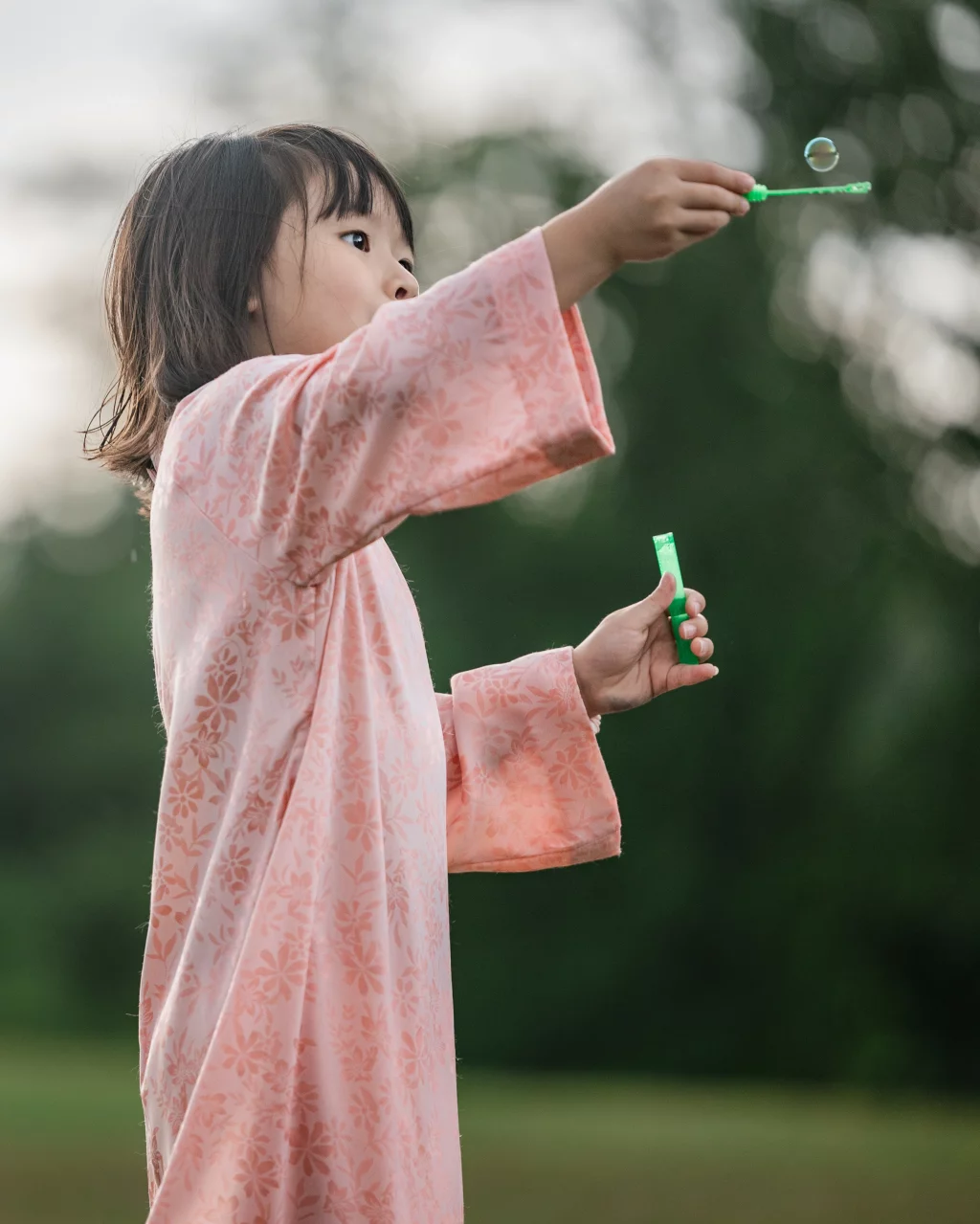 Robbie & Hannah's Wedding @ Edinburgh West, Taylors SC: Child in a pink floral dress blowing a bubble while holding a green bubble wand outdoors.