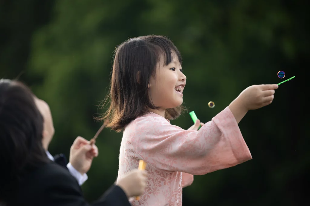 Robbie & Hannah's Wedding @ Edinburgh West, Taylors SC: Smiling child in a pink floral kimono blows bubbles outdoors while an adult reaches toward her.