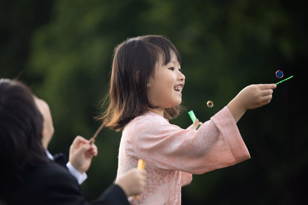 Robbie & Hannah's Wedding @ Edinburgh West, Taylors SC: Smiling child in a pink floral kimono blows bubbles outdoors while an adult reaches toward her.
