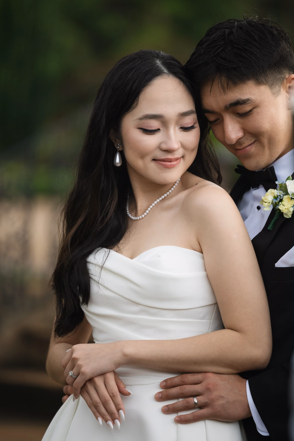 Robbie & Hannah's Wedding @ Edinburgh West, Taylors SC: Bride and groom embrace outdoors, the bride in a strapless white gown and pearls, the groom in a black tuxedo with boutonniere.