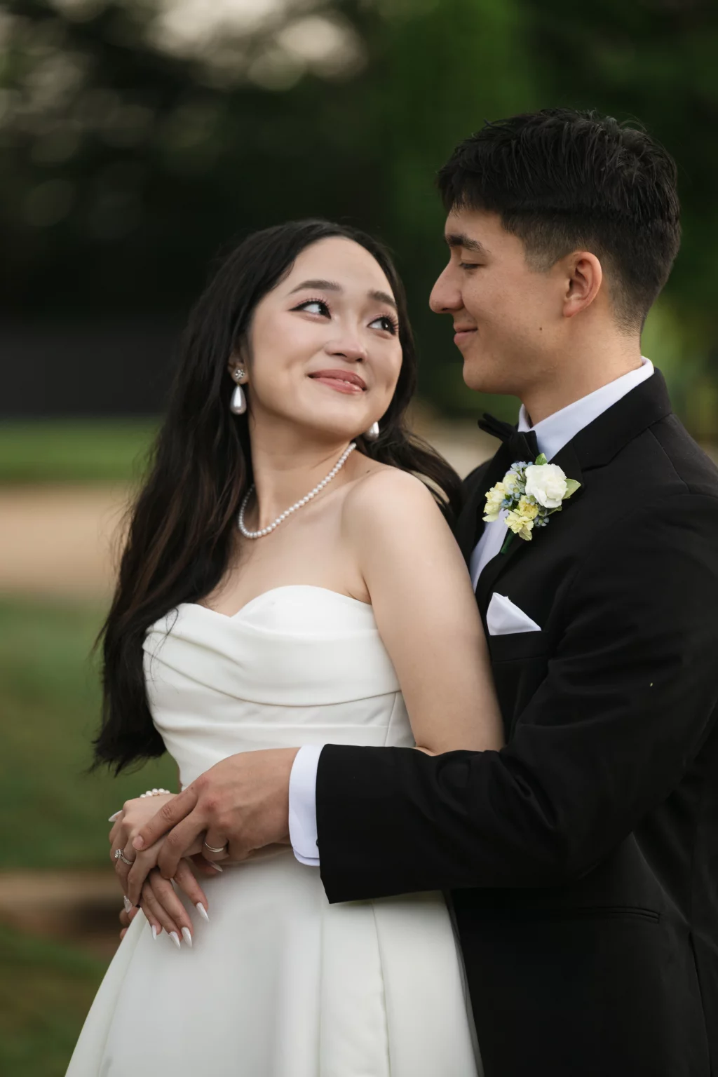 Robbie & Hannah's Wedding @ Edinburgh West, Taylors SC: Bride in a strapless white gown and groom in a black tuxedo embrace outdoors, gazing affectionately at each other.