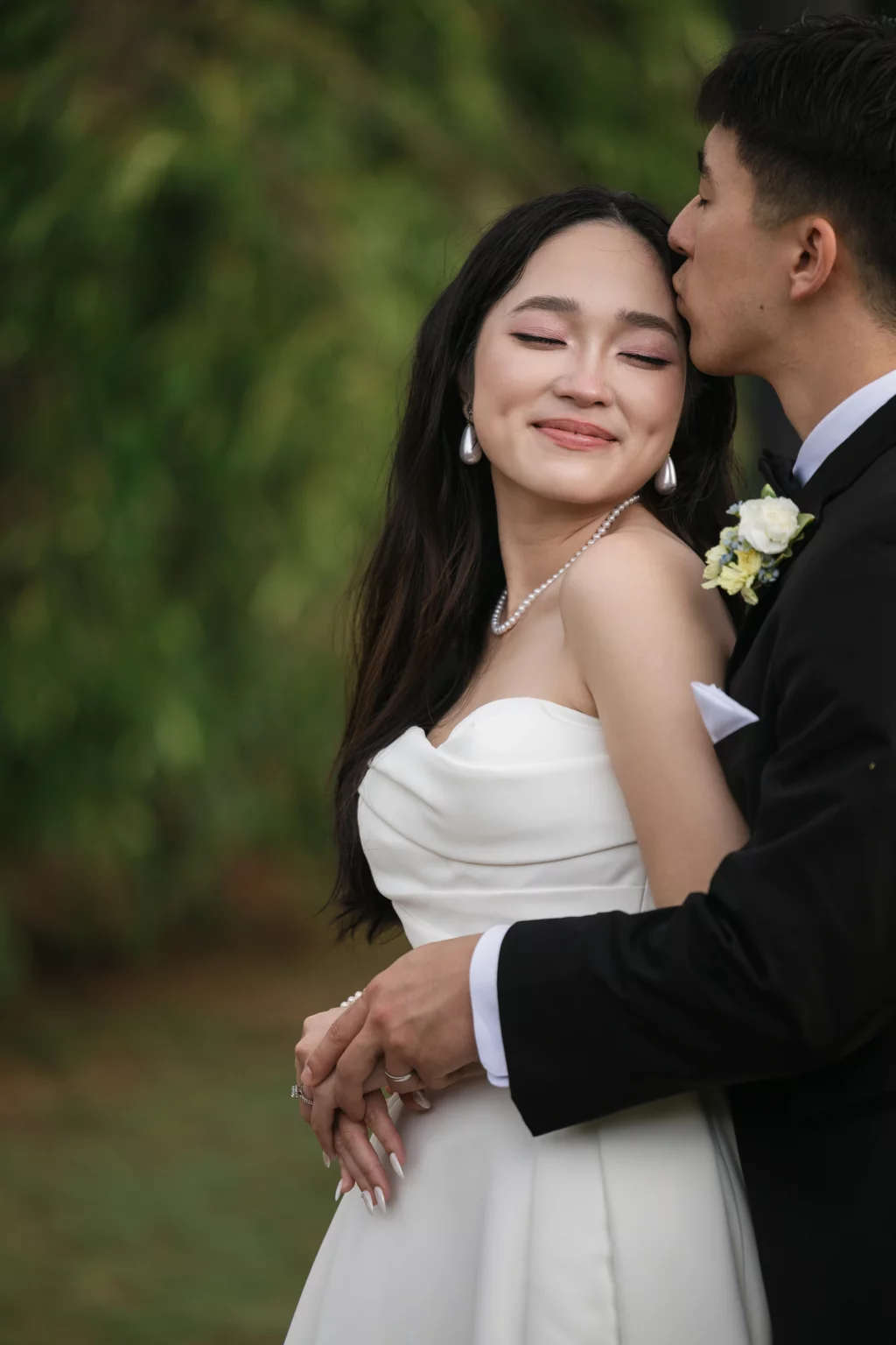 Robbie & Hannah's Wedding @ Edinburgh West, Taylors SC: Bride in a strapless white gown smiles as her groom kisses her forehead outdoors.