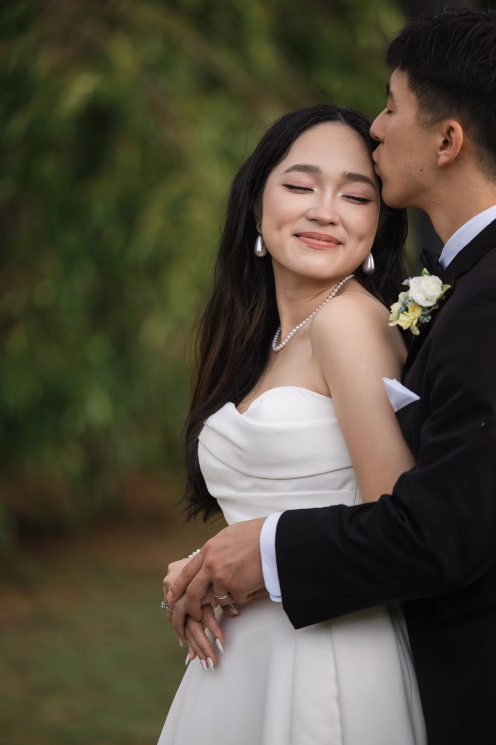 Robbie & Hannah's Wedding @ Edinburgh West, Taylors SC: Bride in a strapless white gown smiles as her groom kisses her forehead outdoors.