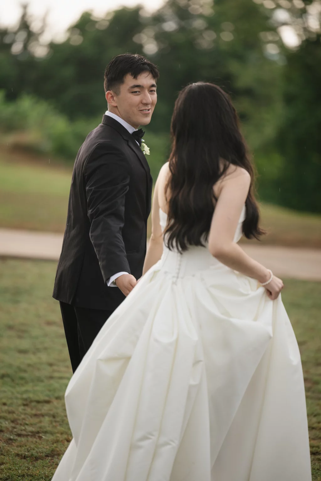 Robbie & Hannah's Wedding @ Edinburgh West, Taylors SC: Groom in a black tuxedo looks back at the bride, who lifts her white wedding dress outdoors.