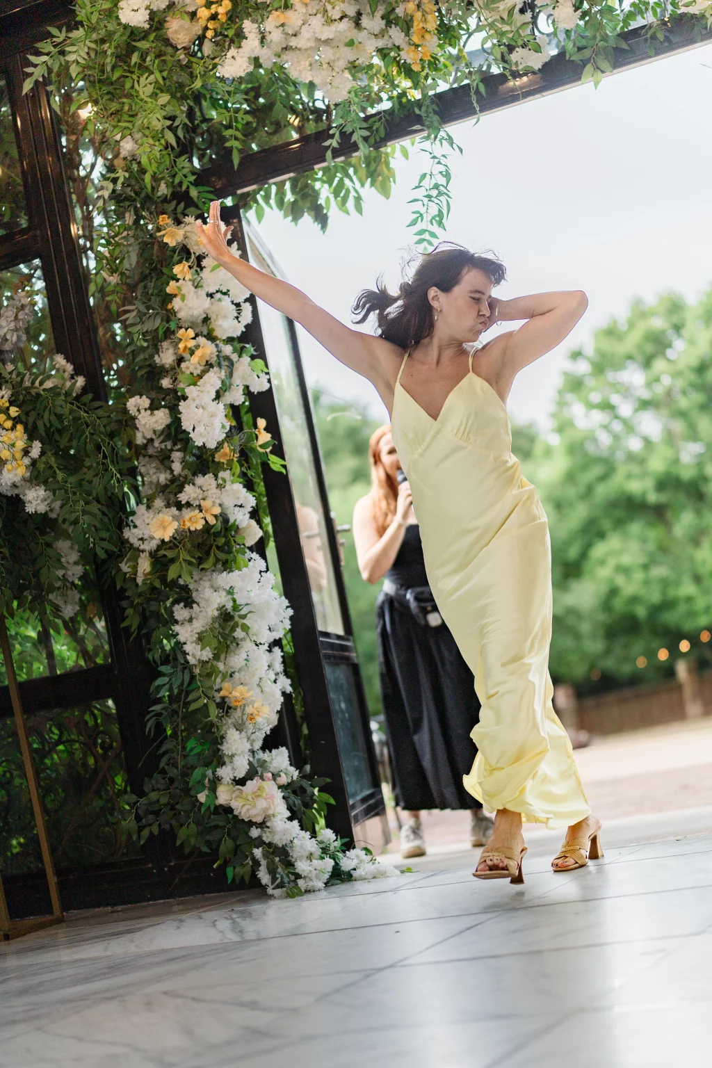 Robbie & Hannah's Wedding @ Edinburgh West, Taylors SC: Woman in a pale yellow dress dances beneath a flower-covered arch at a wedding.