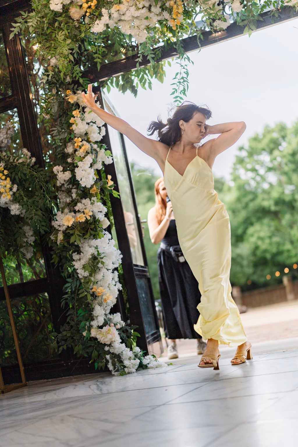 Robbie & Hannah's Wedding @ Edinburgh West, Taylors SC: Woman in a pale yellow dress dances beneath a flower-covered arch at a wedding.