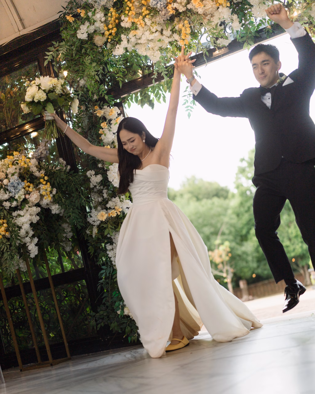 Robbie & Hannah's Wedding @ Edinburgh West, Taylors SC: Bride holding a bouquet and groom in a tuxedo jump beneath a flower-covered arch at their wedding.