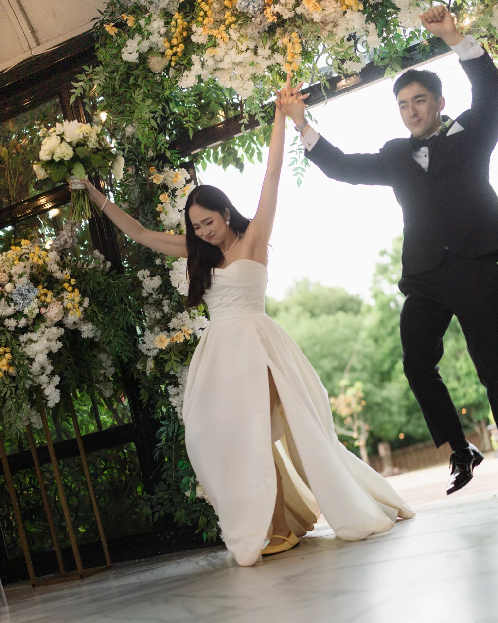 Robbie & Hannah's Wedding @ Edinburgh West, Taylors SC: Bride holding a bouquet and groom in a tuxedo jump beneath a flower-covered arch at their wedding.