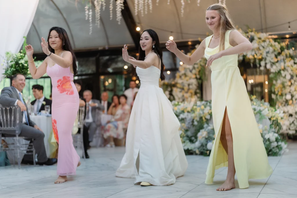 Robbie & Hannah's Wedding @ Edinburgh West, Taylors SC: Three women dance barefoot at a wedding reception, wearing pink, white, and yellow dresses, with guests and floral d&eacute;cor behind them.