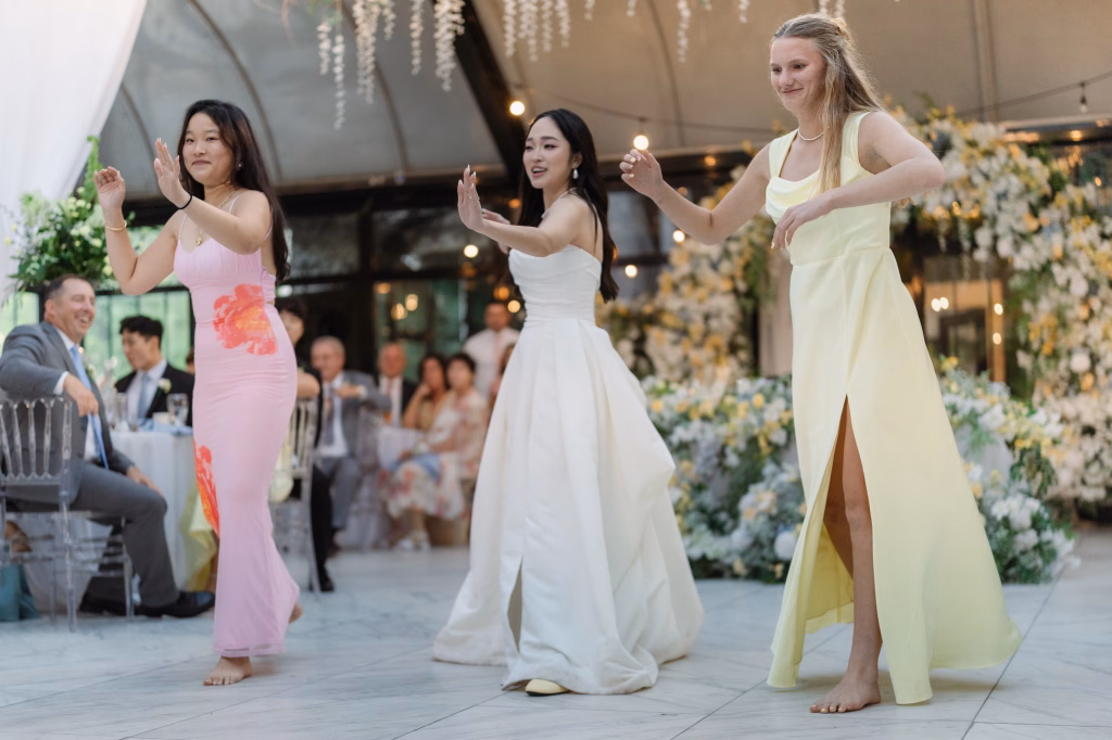 Robbie & Hannah's Wedding @ Edinburgh West, Taylors SC: Three women dance barefoot at a wedding reception, wearing pink, white, and yellow dresses, with guests and floral d&eacute;cor behind them.