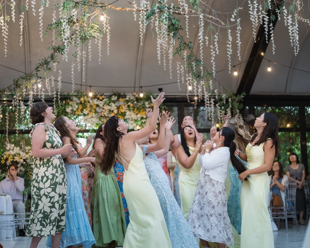 Robbie & Hannah's Wedding @ Edinburgh West, Taylors SC: Wedding guests in colorful dresses reach upward for a bouquet beneath hanging flowers and string lights.