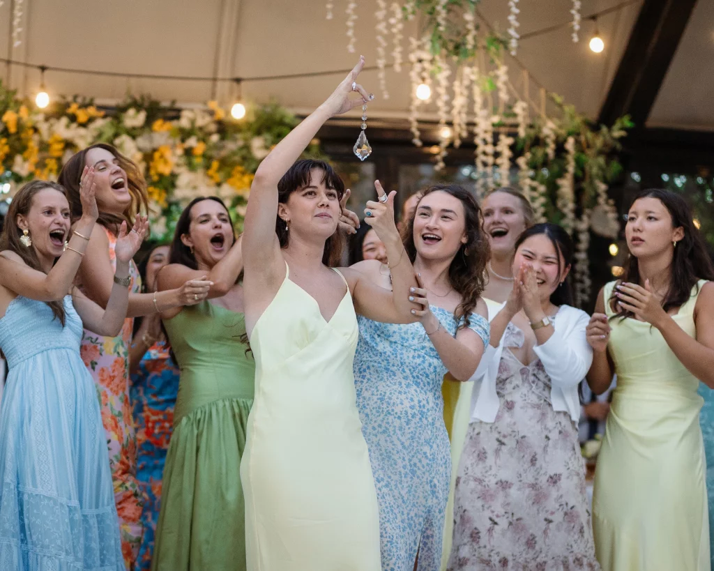 Robbie & Hannah's Wedding @ Edinburgh West, Taylors SC: Excited wedding guests cheer as a woman holds up a crystal ornament beneath floral decorations and string lights.
