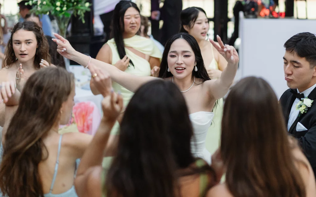 Robbie & Hannah's Wedding @ Edinburgh West, Taylors SC: Bride in a white dress dances with raised arms among wedding guests, beside a groom in a black tuxedo.