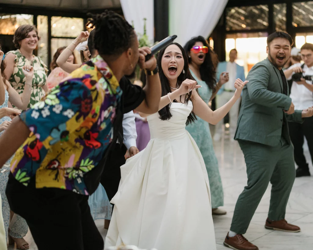 Robbie & Hannah's Wedding @ Edinburgh West, Taylors SC: Bride and wedding guests dance energetically indoors while a singer performs with a microphone.