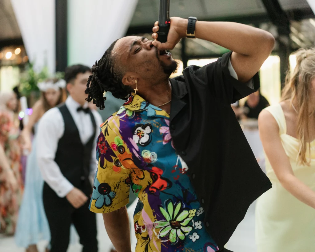 Robbie & Hannah's Wedding @ Edinburgh West, Taylors SC: Guest sings into a microphone while dancing at a wedding reception, wearing a colorful patterned shirt and black vest.